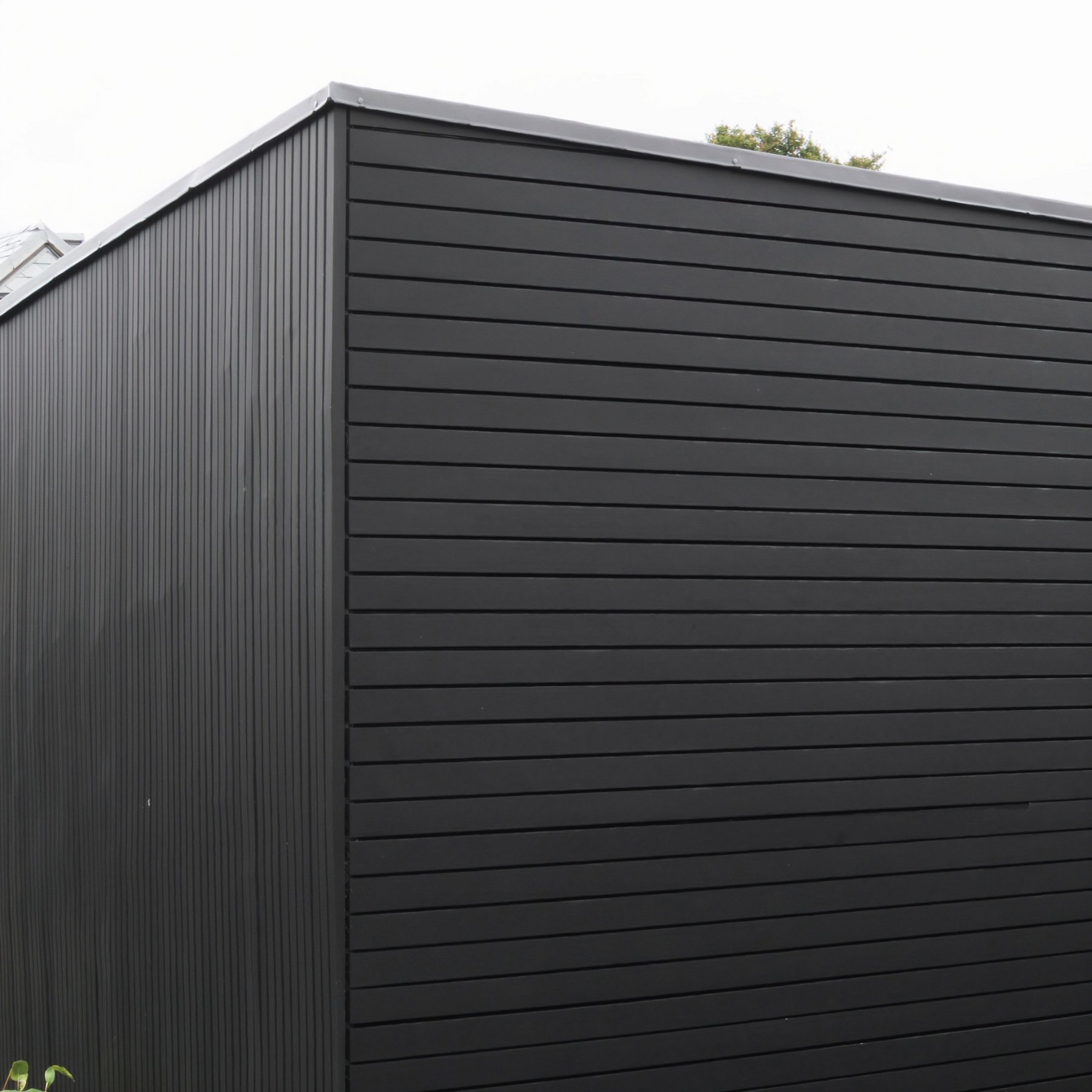 External corner of a garden room where vertical ribbed metal cladding meets horizontal composite boards, with a slim coping along the top edge